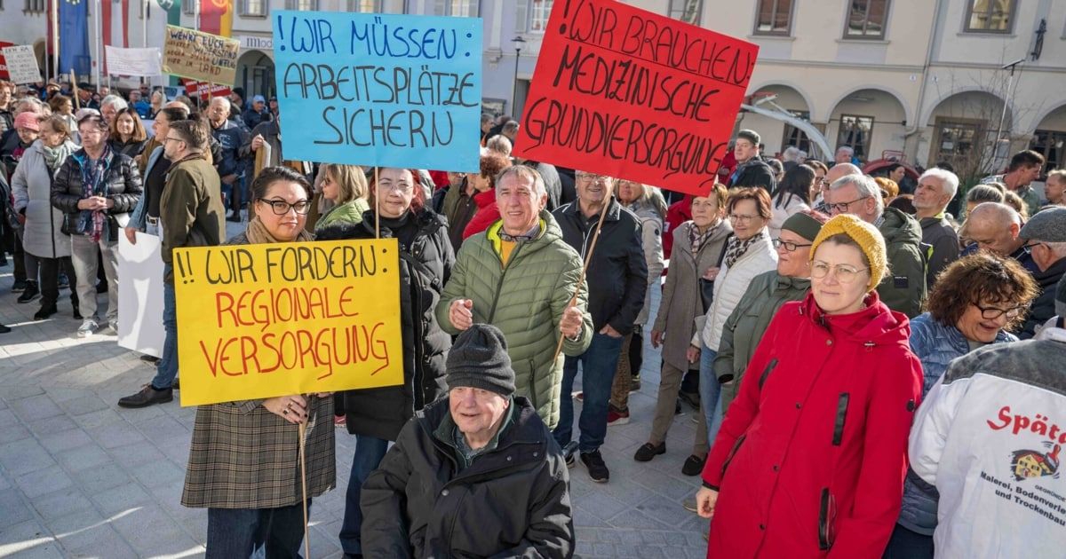 Demonstranten halten Schilder für medizinische Versorgung und Arbeitsplatzsicherheit. Eine vielfältige Menschenmenge versammelt sich, einige im Rollstuhl, tragen Mützen und Brillen.