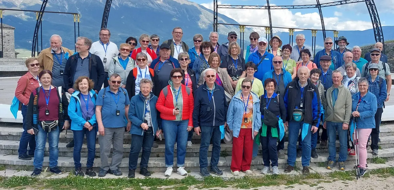 Eine Gruppe von Menschen mit Sonnenbrillen und Namensschildern posiert auf einer Treppe mit Bergen im Hintergrund für ein Foto.