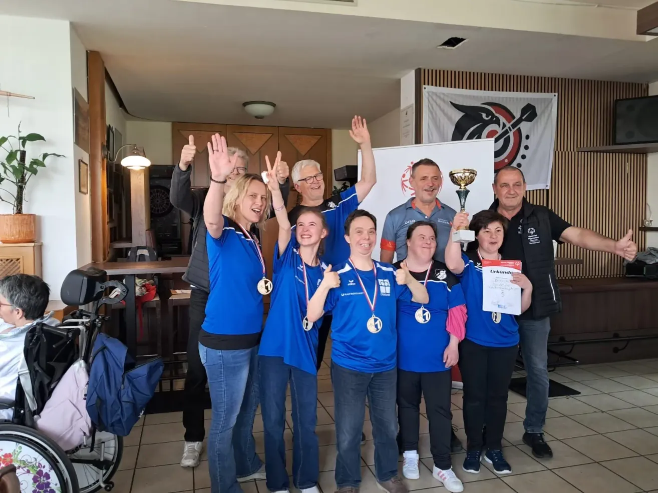 Eine Gruppe von Personen in blauen Trikots und mit Medaillen feiert vor einem Banner, während sie Trophäen und Urkunden halten. Sie werden von einem lächelnden Mann mit einer Trophäe und einem anderen Mann mit einer Urkunde umgeben.