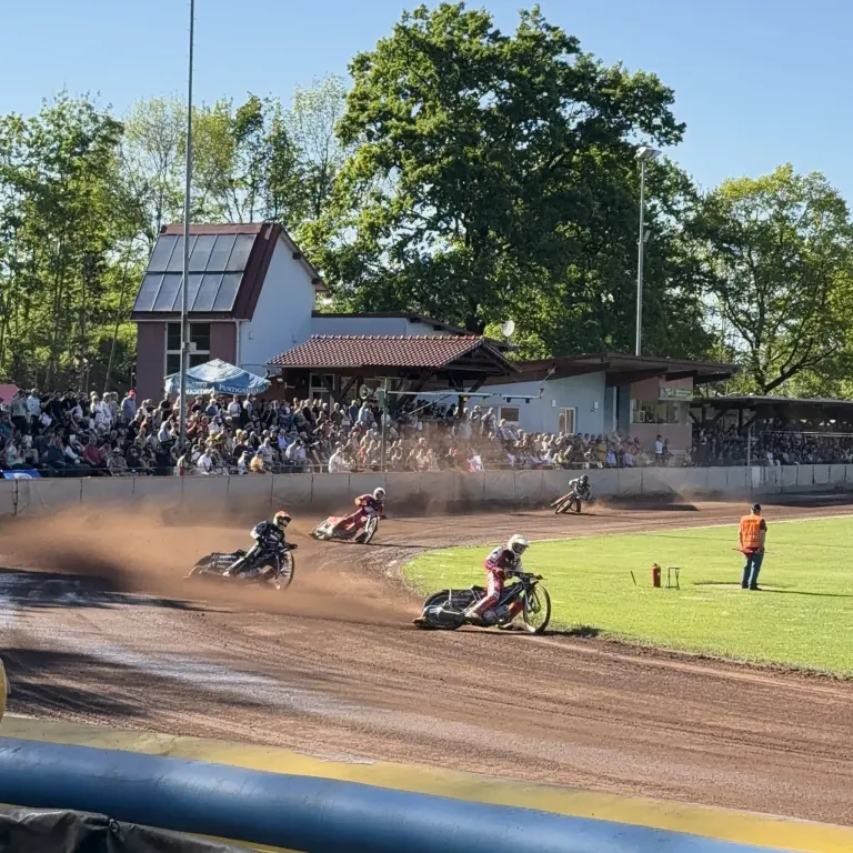Fahrer mit Helmen konkurrieren auf einer Dirt-Bahn, während ein Publikum aus den Tribünen zuschaut. Es gibt mehrere Motorräder und Bäume im Hintergrund.