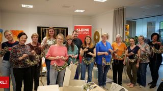 Eine Gruppe von Frauen posiert für ein Foto mit Kränzen in einer Workshop-Raum mit einem roten Banner, das Kleine Eitgung liest.