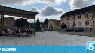 Ein sonniger Tag im Barista's Café, wo Menschen unter einem Vordach sitzen. Gebäude und geparkte Autos sind im Hintergrund.