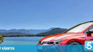 Ein rotes Auto in der Nähe eines Sees geparkt, Berge im Hintergrund, klarer blauer Himmel darüber.