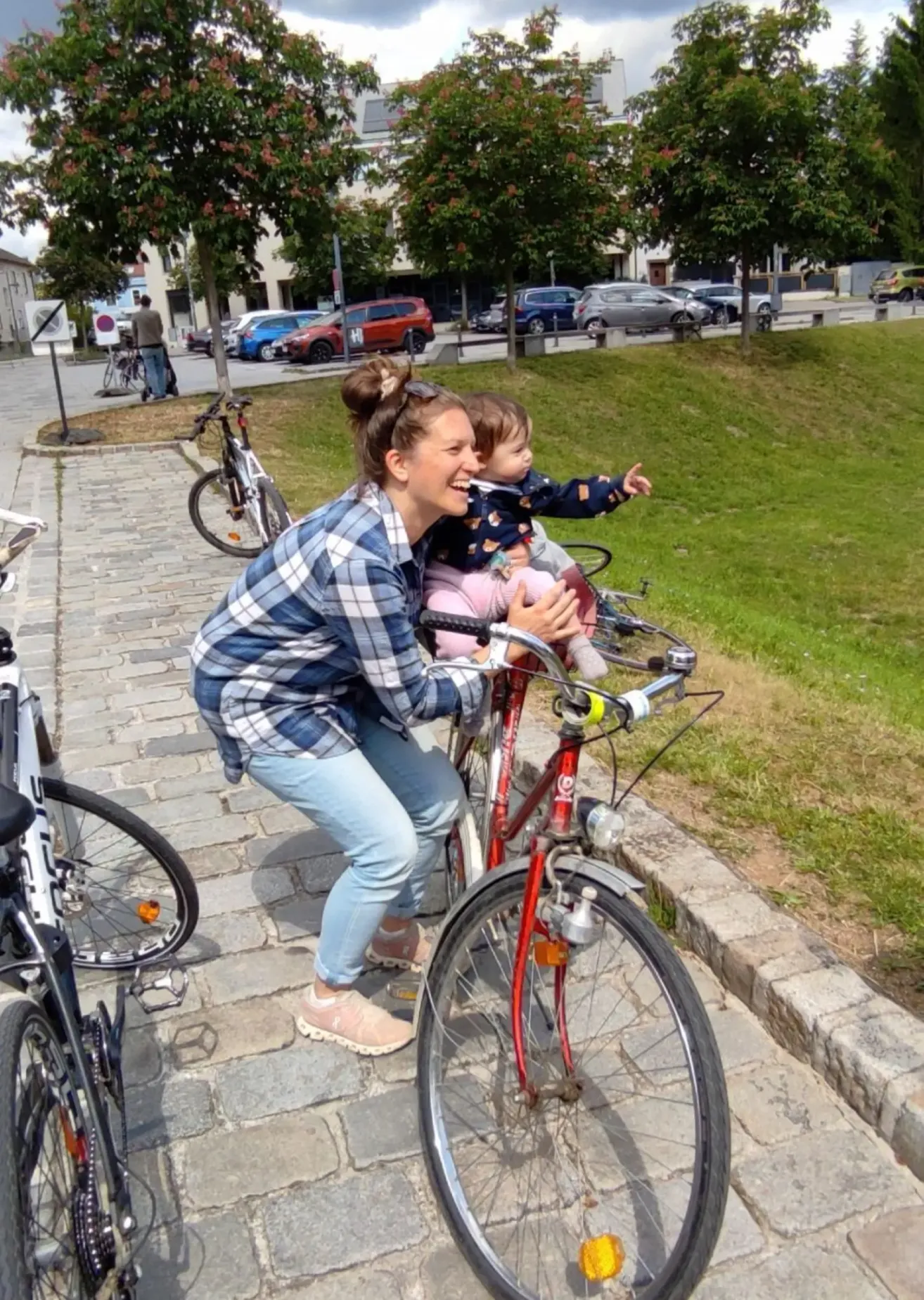 Eine Frau in einem karierten Hemd fährt mit einem Baby auf einem Fahrrad an einem sonnigen Tag auf einem Kopfsteinpflasterweg mit geparkten Autos und Grün im Hintergrund.