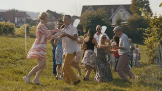 Eine Gruppe von Menschen tanzt auf einer Grasfläche mit Häusern im Hintergrund. Einige tragen Brillen und eine Person hat eine Armbanduhr.