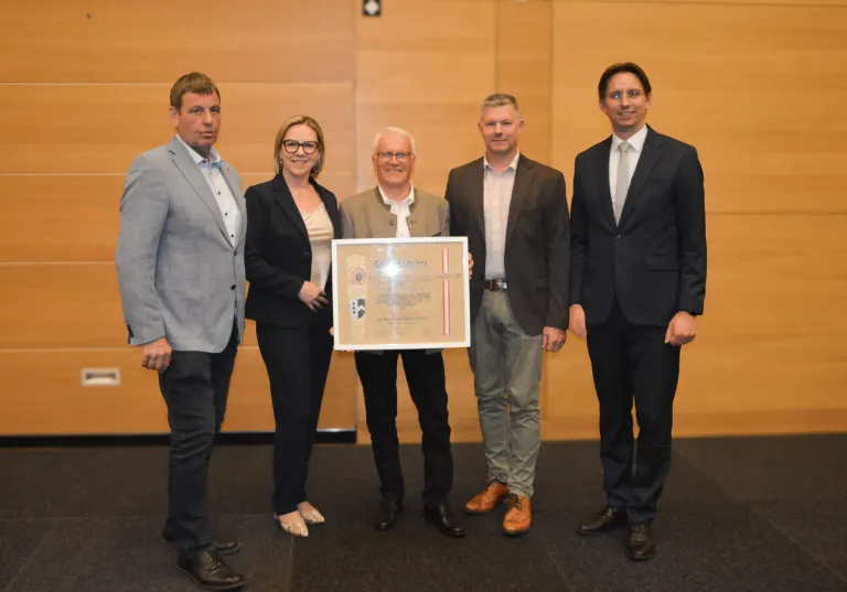 Fünf Personen stehen für ein Foto; einer hält eine gerahmte Auszeichnung. Der Mann in der Mitte trägt einen beigen Anzug. Die anderen vier sind in schwarzen Anzügen. Sie befinden sich in einem Konferenzraum.