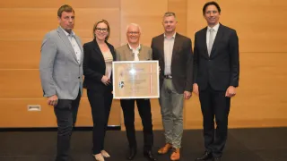 Fünf Personen stehen für ein Foto; einer hält eine gerahmte Auszeichnung. Der Mann in der Mitte trägt einen beigen Anzug. Die anderen vier sind in schwarzen Anzügen. Sie befinden sich in einem Konferenzraum.