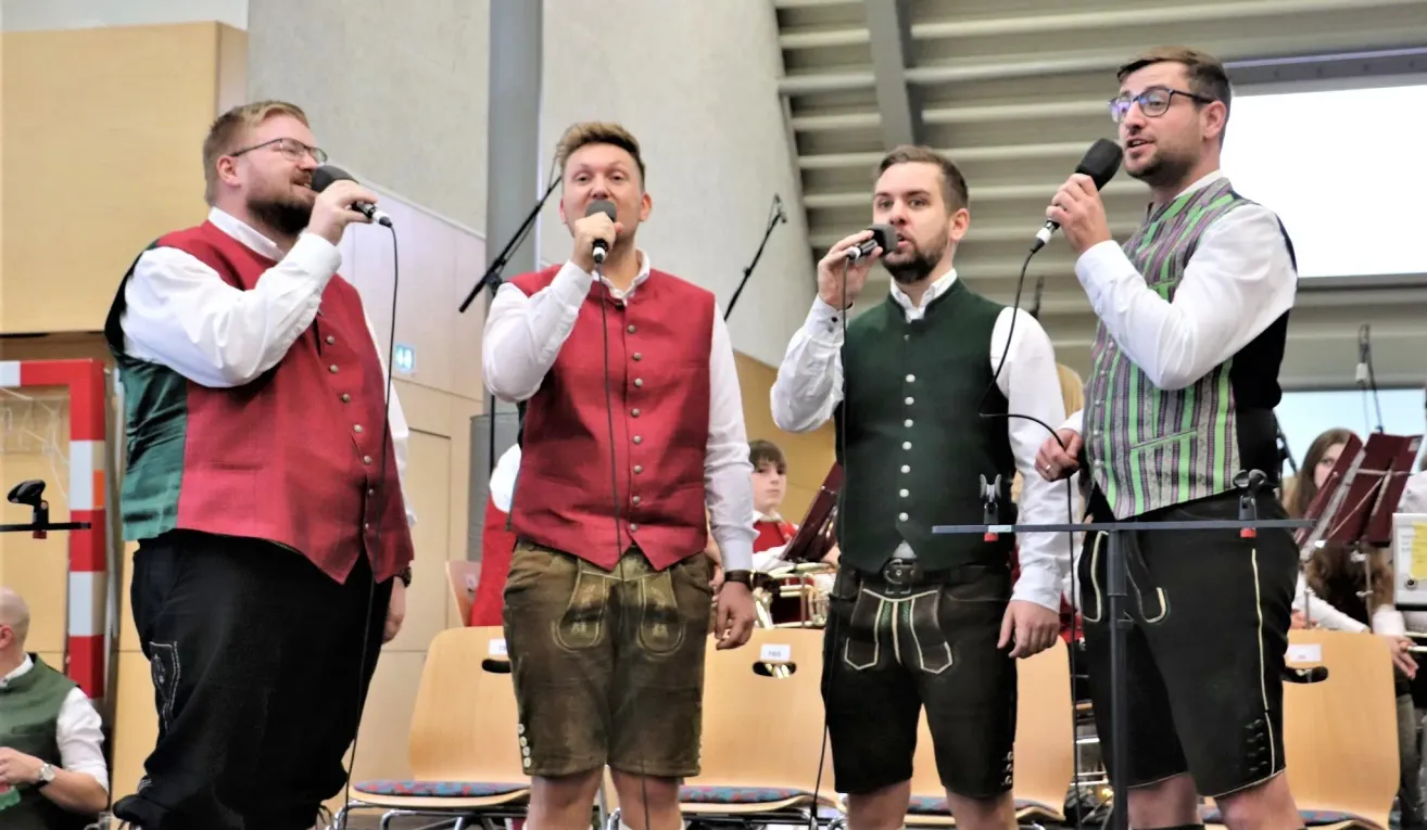 Vier Männer in traditioneller deutscher Kleidung, einschließlich Lederhosen, singen in Mikrofone auf einer Bühne. Eine Band und Publikum sind hinter ihnen.