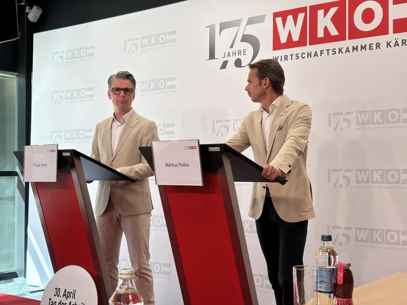 Zwei Männer stehen an Rednerpulten vor einem Banner, das '175 WK' steht. Der Mann auf der linken Seite trägt eine Brille und einen hellen Anzug, während der Mann auf der rechten Seite einen beigen Blazer trägt. Es gibt Flaschen und ein Glas auf dem Rednerpult.