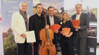 Fünf Personen posieren für ein Foto in einem Raum mit Holzboden. Sie halten rote Broschüren und ein Cello. Sie stehen neben Bannern.