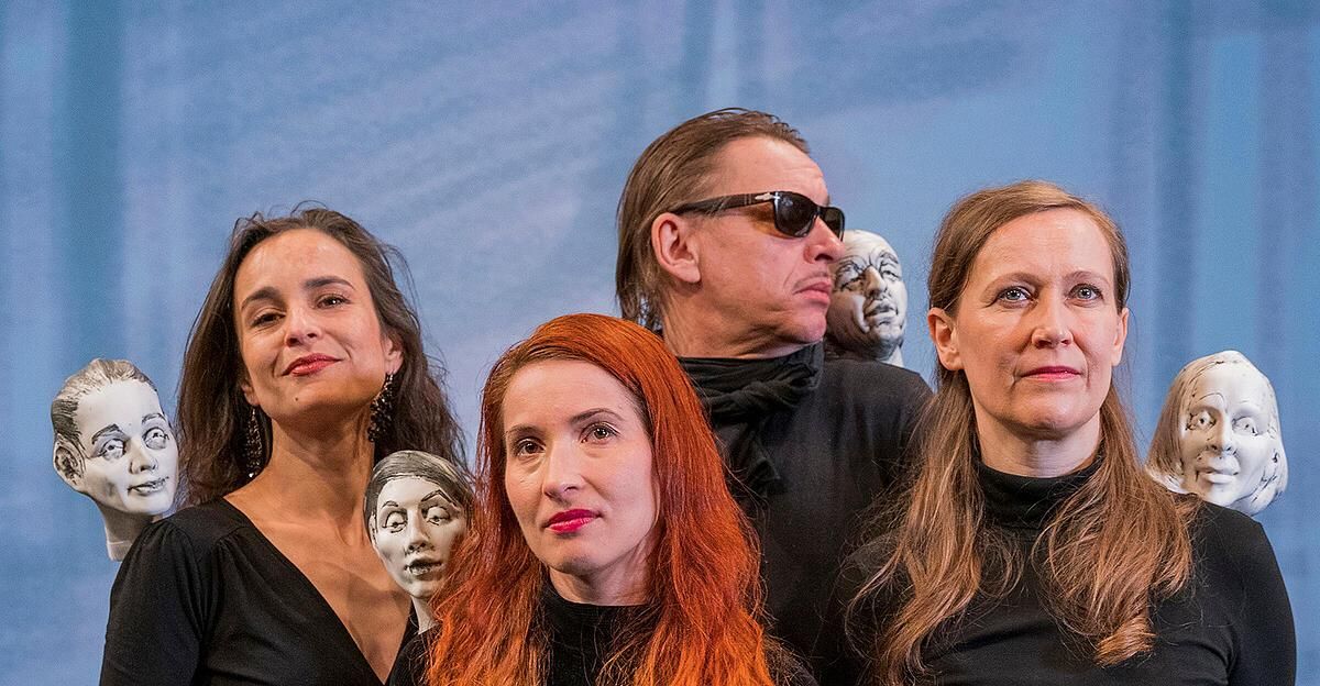 Fünf Personen, drei Frauen und zwei Männer, stehen eng beieinander und lächeln für ein Foto. Dahinter befinden sich zwei Kopfskulpturen, von denen eine auf der Schulter eines Mannes ist.