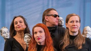 Fünf Personen, drei Frauen und zwei Männer, stehen eng beieinander und lächeln für ein Foto. Dahinter befinden sich zwei Kopfskulpturen, von denen eine auf der Schulter eines Mannes ist.