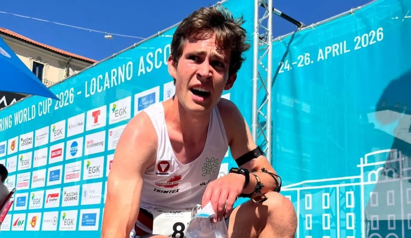 Ein Läufer sitzt mit offenem Mund auf dem Boden und hält eine kleine Plastiktüte. Er trägt ein weißes Shirt mit der Nummer 8. Hinter ihm steht ein blauer Banner mit der Aufschrift 'Locarno Asco'.