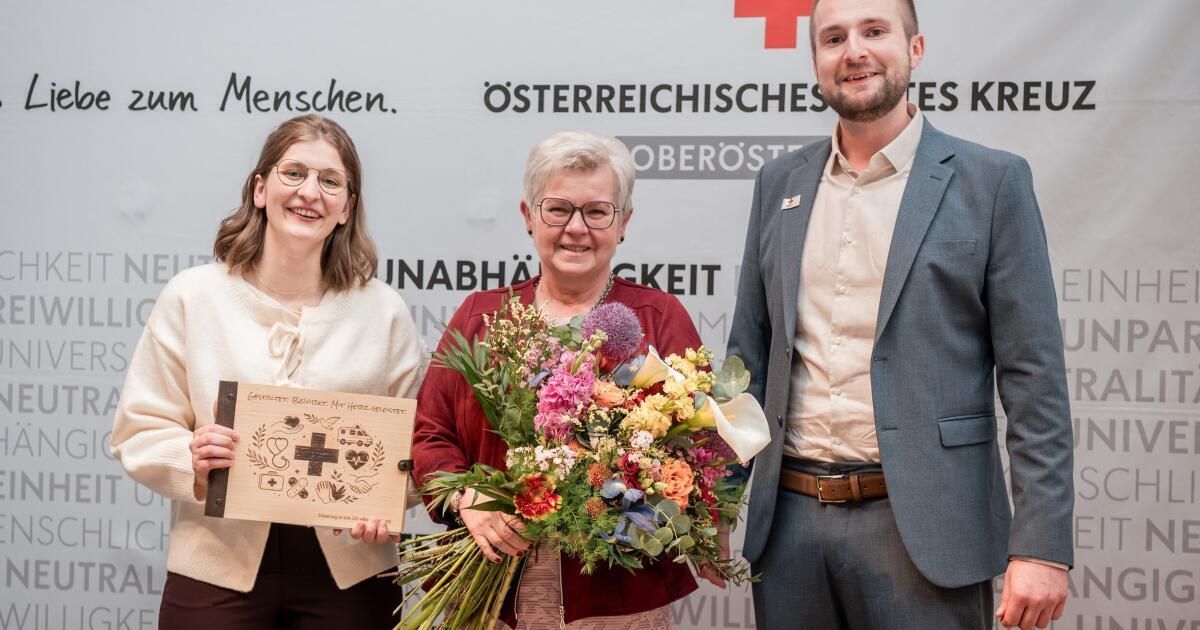 Drei Menschen stehen lächelnd vor einem Banner. Die Frau auf der linken Seite hält eine Plakette, die Frau in der Mitte hält Blumen und der Mann auf der rechten Seite trägt einen Anzug.