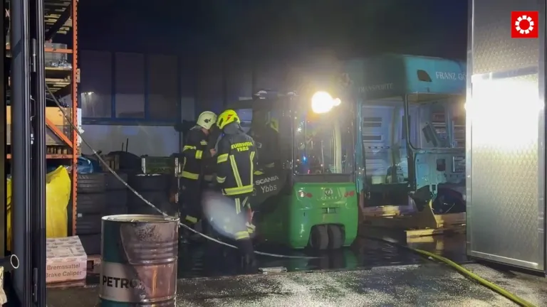 Zwei Feuerwehrleute in gelben und schwarzen Uniformen arbeiten an einem grünen Nutzfahrzeug mit hellem Licht, das in einem Lagerhaus geparkt ist.