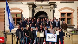 Eine Gruppe von Männern und Frauen in passenden blauen Uniformen steht vor einem Gebäude mit einer Flagge, hält Medaillen und Trophäen. Sie werden von anderen in formeller Kleidung begleitet. Ein Schild mit der Aufschrift Hartberg wird von einem der Männer gehalten.