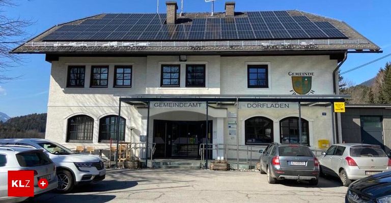 Ein weißes Gebäude mit Solarpanelen auf dem Dach, beschriftet als Gemeindeamt Dorfladen. Zwei Autos sind vor dem Gebäude geparkt.