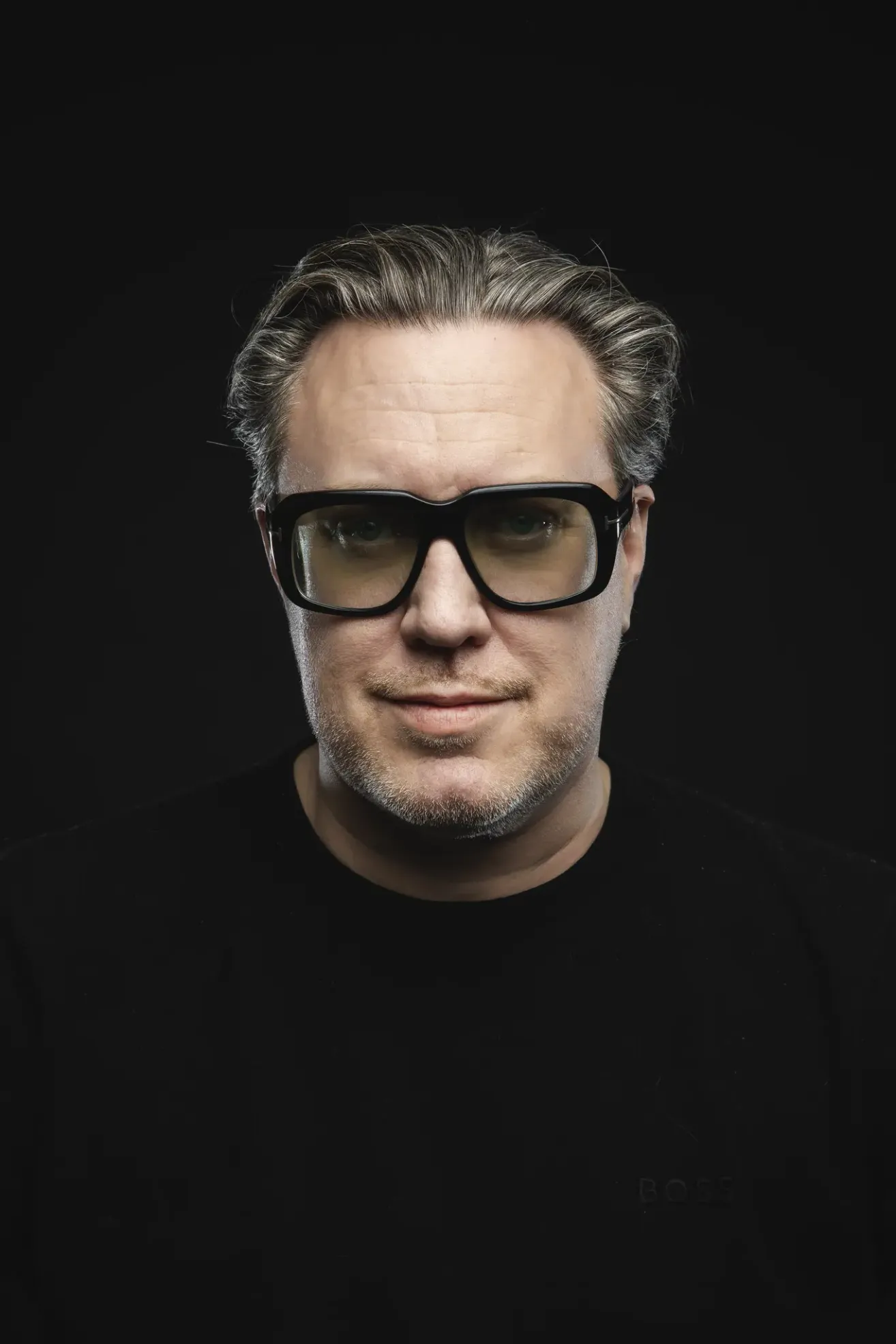 Ein Mann mit grauem Haar, der eine Brille und ein schwarzes T-Shirt trägt, lächelt mit einem schwarzen Hintergrund vor sich in die Kamera.