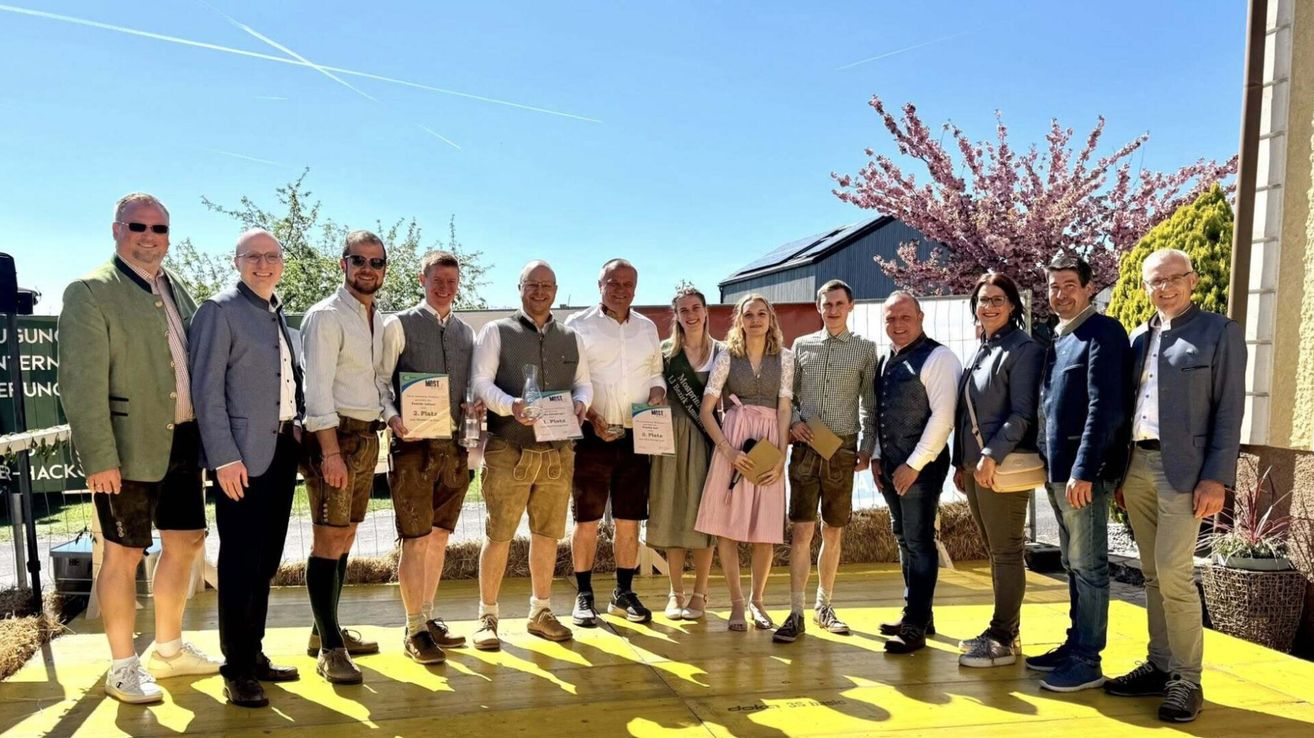Gruppe von Personen auf einer Bühne, in traditioneller Kleidung. Sie halten Urkunden und Auszeichnungen. Dahinter befindet sich eine Kulisse aus Gebäuden und Bäumen.