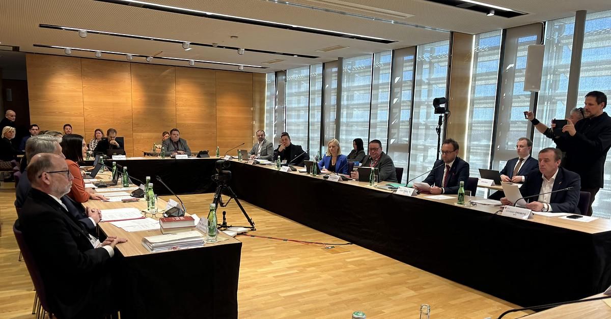 Eine Besprechung mit mehreren Personen, die an einem langen Tisch sitzen, mit Mikrofonen und Flaschen. Der Raum hat Holzwände und Glasfenster.