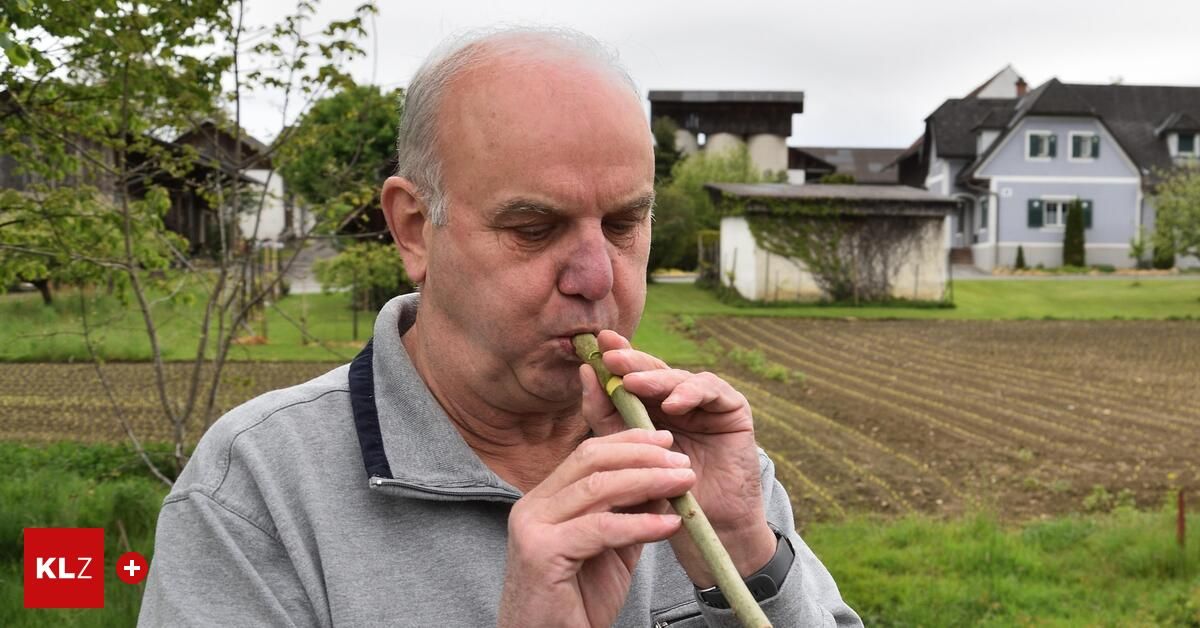 Ein älterer Mann spielt eine Holzflöte im Freien auf einem Feld mit Häusern im Hintergrund.