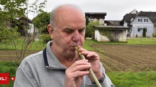 Ein älterer Mann spielt eine Holzflöte im Freien auf einem Feld mit Häusern im Hintergrund.
