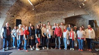 Eine Gruppe von Menschen in verschiedenen Outfits steht in einem Raum mit Backsteinwänden, einige halten Papiere in der Hand, wahrscheinlich für ein Foto posend.