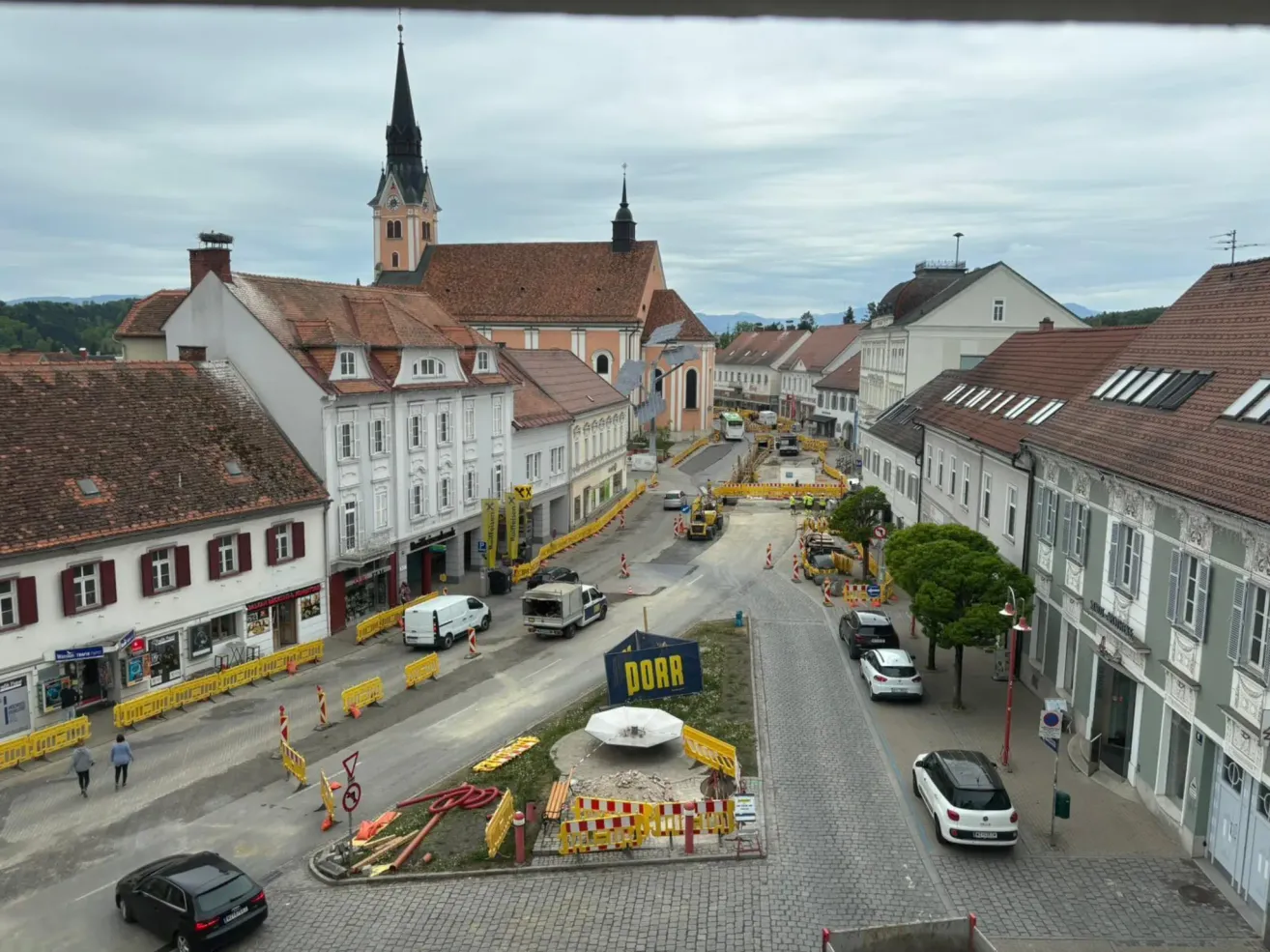 Ein Ortszentrum mit laufenden Bauarbeiten. Gebäude, Bauzäune und Fahrzeuge sind zu sehen. Die Straße ist teilweise blockiert mit einem blauen Schild, das 'DORR' steht.