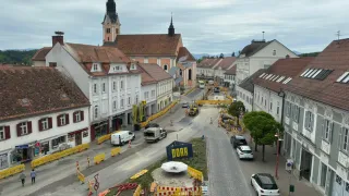 Ein Ortszentrum mit laufenden Bauarbeiten. Gebäude, Bauzäune und Fahrzeuge sind zu sehen. Die Straße ist teilweise blockiert mit einem blauen Schild, das 'DORR' steht.