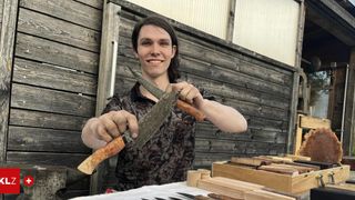Eine Person hält ein Messer in beiden Händen vor einem Holztisch und einer Holzwand.