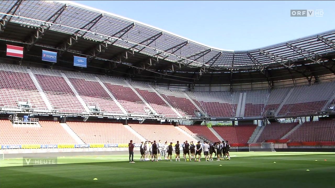 Eine Gruppe von Spielern trainiert auf einem Fußballfeld, mit leeren roten Sitzen im Hintergrund. Ein blaues Schild befindet sich in der oberen Reihe des Stadions.