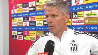 Ein Mann in einem weißen und schwarzen Trikot mit LASK Logo spricht in ein Mikrofon und steht vor einer Wand mit verschiedenen Werbelogos.