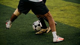 Ein Fußballspieler steht auf einem Feld und ist dabei, einen Ball mit dem Fuß zu schießen. Der Ball hat ein schwarzes und weißes Muster. Das Bein des Spielers ist in der Luft.