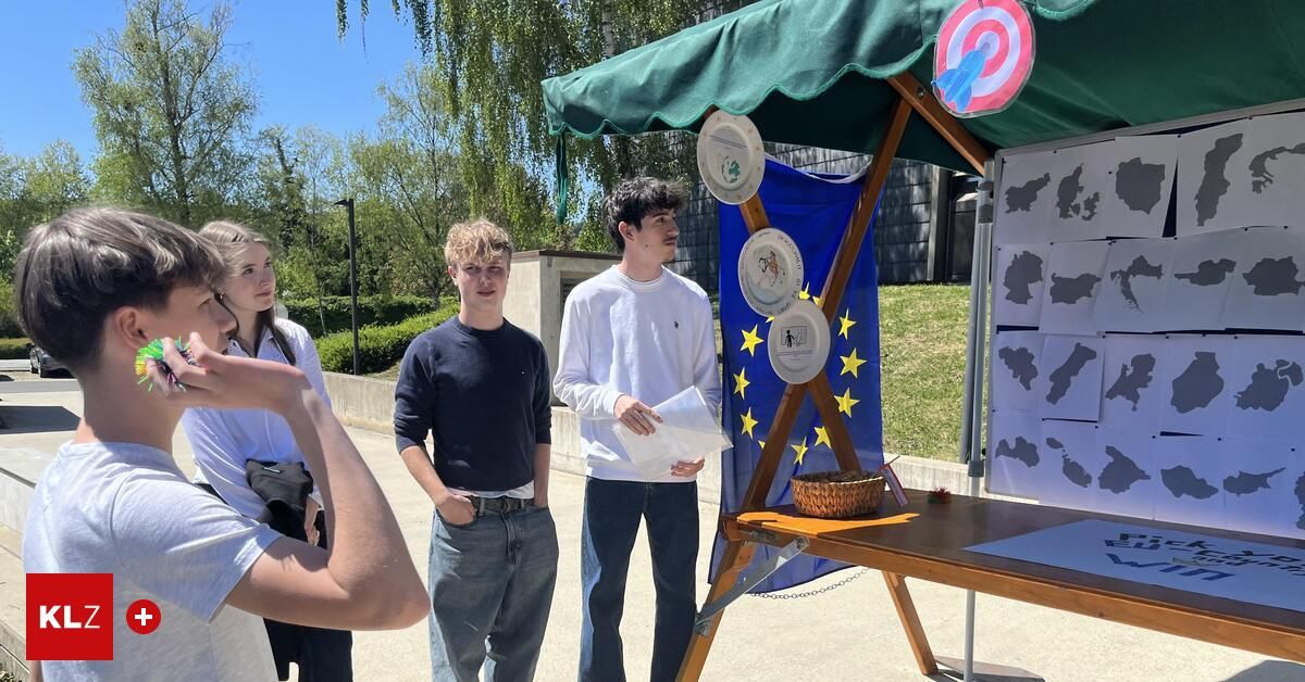 Drei junge Leute stehen an einem Stand mit einer Europaflagge und verschiedenen Objekten und Tellern. Eine Person hält ein Papier und spricht.