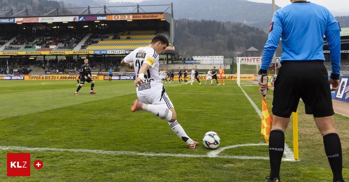 Ein Fußballspieler tritt den Ball in der Nähe der Torlinie. Ein Schiedsrichter hält eine Flagge, und andere Spieler rennen auf dem Feld. Berge sind im Hintergrund.