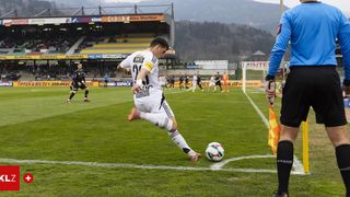 Ein Fußballspieler tritt den Ball in der Nähe der Torlinie. Ein Schiedsrichter hält eine Flagge, und andere Spieler rennen auf dem Feld. Berge sind im Hintergrund.
