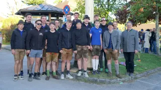 Eine Gruppe junger Männer und Erwachsener posiert für ein Foto in traditioneller deutscher Kleidung, einige tragen Hüte und Brillen. Sie stehen vor einem Pfosten und Gras, mit einem Gebäude, Bäumen und einem Schild im Hintergrund.