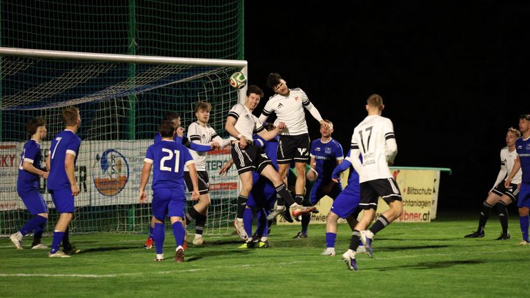 Fußballspieler in blauen Trikots kämpfen vor dem Tor, ein Spieler springt, um den Ball zu köpfen, während andere um die Kontrolle ringen.