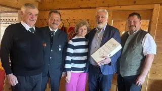 Fünf Menschen stehen in einem Raum mit Holzwänden und lächeln. Der Mann in der Mitte hält eine Box. Die Frau neben ihm trägt ein gestreiftes Shirt und pinke Hose. Die anderen tragen formelle Kleidung.