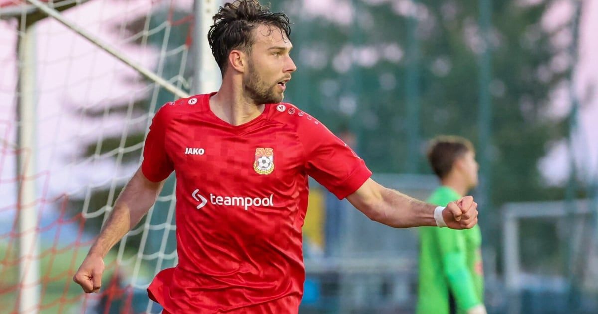 Ein Fußballspieler in einem roten Trikot feiert ein Tor in der Nähe des Netzes, mit einem unscharfen Hintergrund eines anderen Spielers und Bäumen.