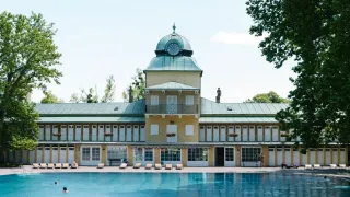 Ein großes gelbes Gebäude mit grünem Dach und Uhrturm steht neben einem großen Swimmingpool. Ein paar Leute liegen auf der Terrasse.