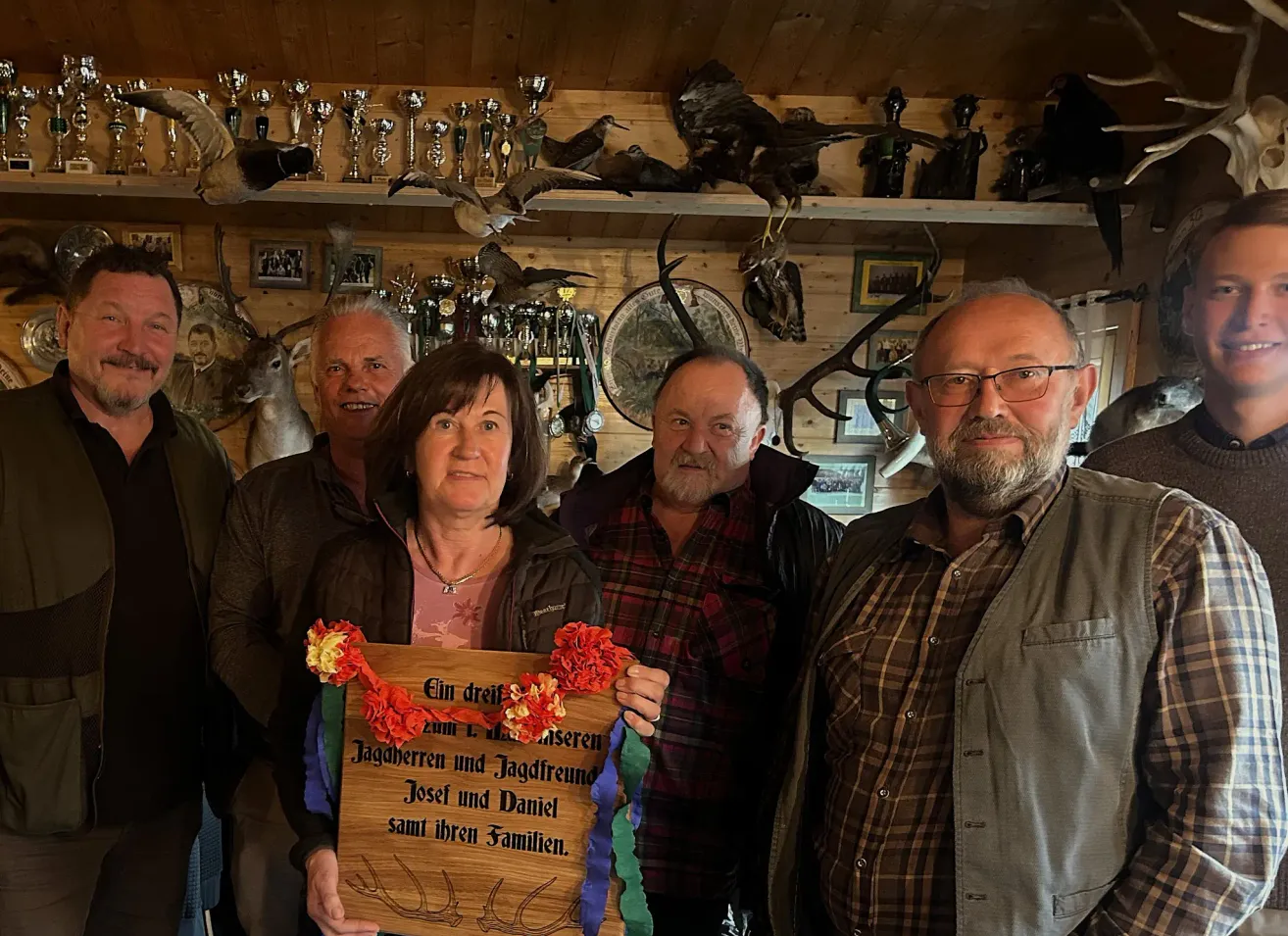 Fünf Menschen stehen in einem Raum mit Trophäen und Tierfiguren. Eine Frau hält eine Holzplakette mit Blumen und Text, die Jagdfreunde ehrt.