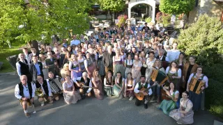 Eine große Gruppe von Menschen in traditioneller Kleidung posiert im Freien für ein Foto und hält Musikinstrumente in den Händen. Sie sind von viel Grün und einem Gebäude mit dem Namen 'Retter' umgeben.