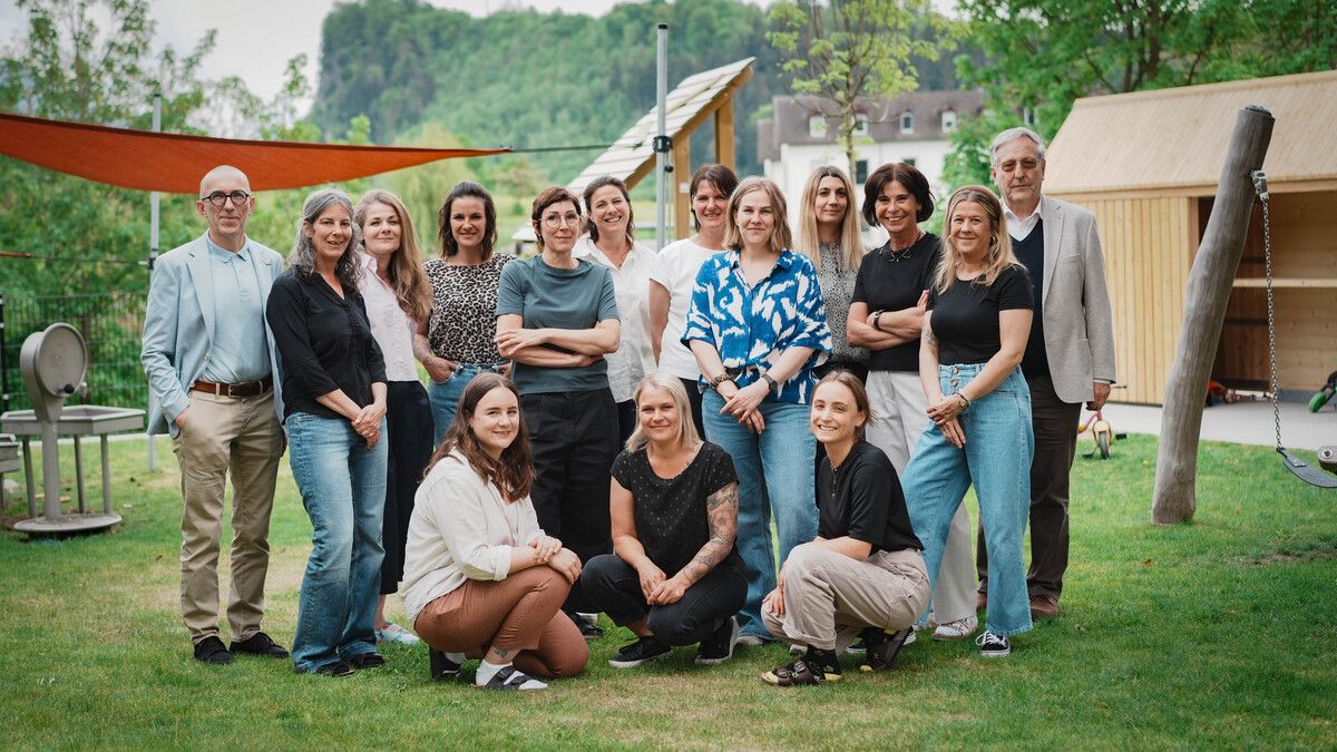 Eine Gruppe von Erwachsenen, sowohl Männer als auch Frauen, posiert für ein Foto auf einem Rasen. Einige knien, andere stehen mit verschränkten Armen. Bäume und ein Gebäude sind im Hintergrund zu sehen.
