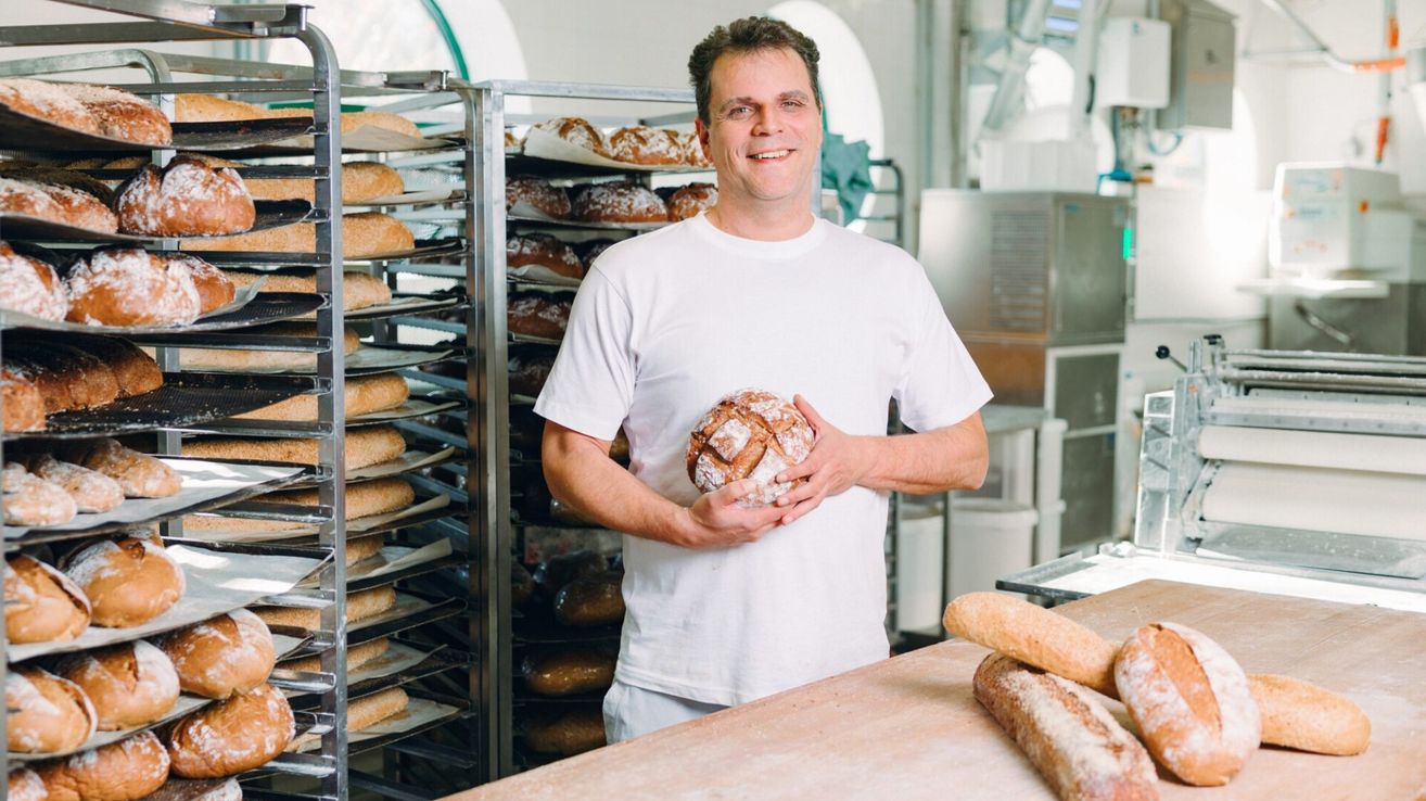 Ein Mann in einem weißen T-Shirt steht in einer Bäckerei, hält ein Brot in den Händen, mit Regalen voller Brot dahinter und einem Tisch mit Brot davor.