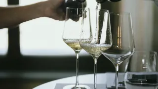 Eine Hand gießt Wein in drei Weinglaser auf einem Tisch. Der Wein ist golden und die Gläser sind klar.