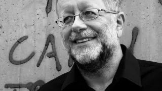 Ein Mann mit Brille, Bart und Lächeln steht vor einer Wand mit Graffiti. Er trägt ein schwarzes Hemd.