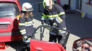Zwei Feuerwehrleute in Schutzkleidung öffnen eine Autotür. Eine Frau steht neben ihnen und beobachtet. Das Auto ist rot und beschädigt.