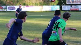 Zwei Spieler in blauen und grünen Trikots sind in einem Fußballspiel engagiert. Ein roter und weißer Fußball ist in der Luft. Ein Mann in einem blauen Hemd steht in der Nähe.