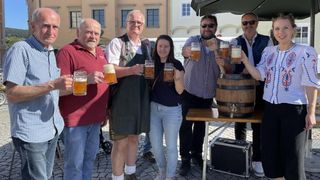 Eine Gruppe von Menschen hält Biergläser vor einem Fass bei einer Outdoor-Veranstaltung und posiert lächelnd für ein Foto.